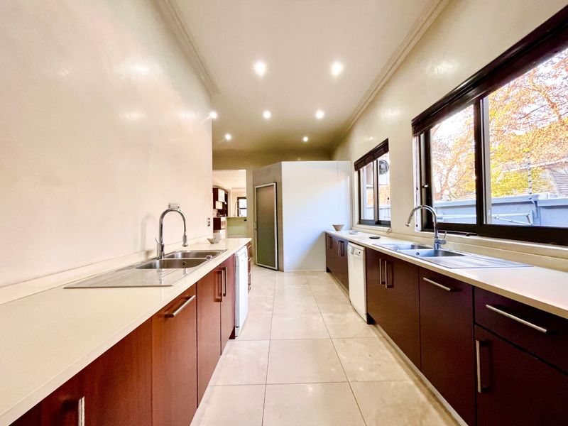 A spacious galley-style kitchen featuring dual stainless steel sinks, dark wood cabinetry, light-colored countertops, and tiled flooring with recessed ceiling lighting.