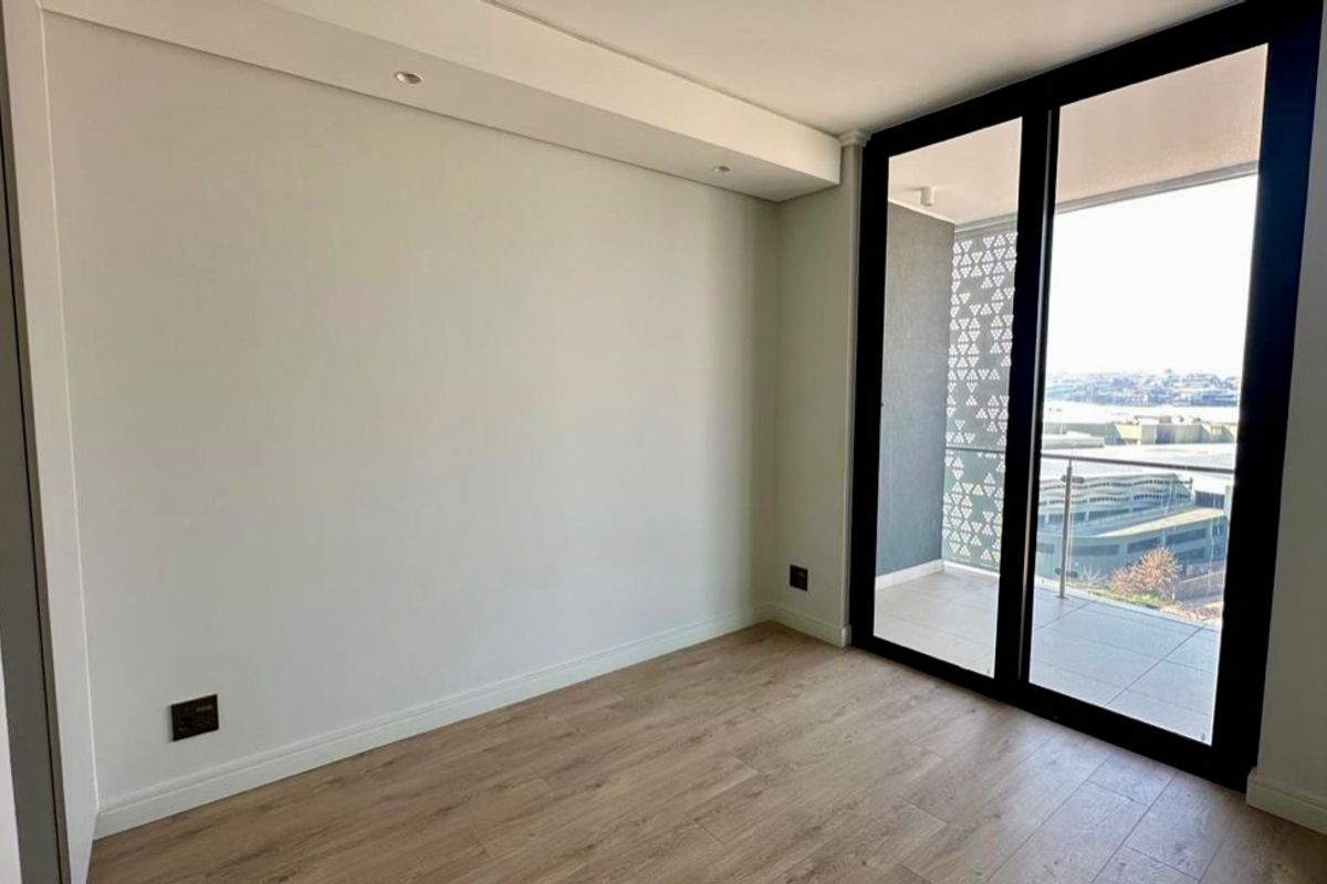 A bright, empty bedroom featuring modern laminate flooring, recessed ceiling lighting, and a large sliding glass door providing access to a private balcony.