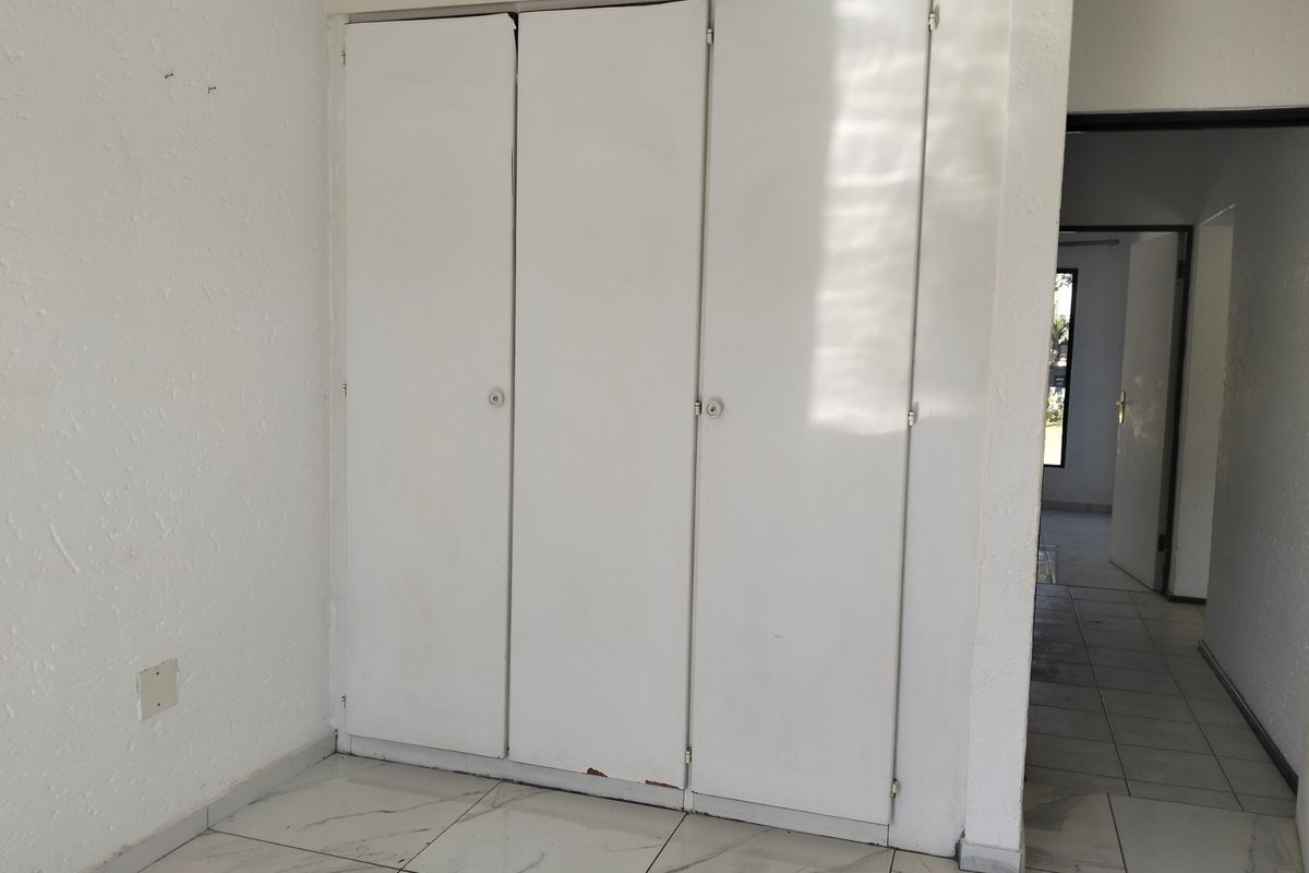 A bedroom interior featuring built-in white wardrobes with tiled flooring, leading into an adjacent hallway area.