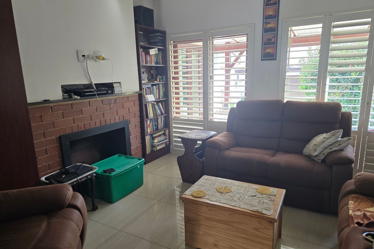 Living room featuring a brick fireplace, tiled flooring, and white plantation shutters providing natural light and access to the outdoor area.