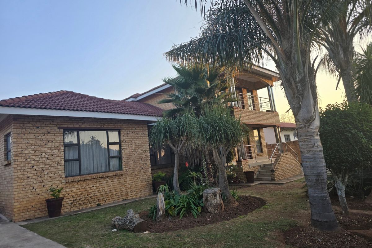 A multi-level face-brick house featuring a tiled roof, balcony, and established garden with mature palm trees.
