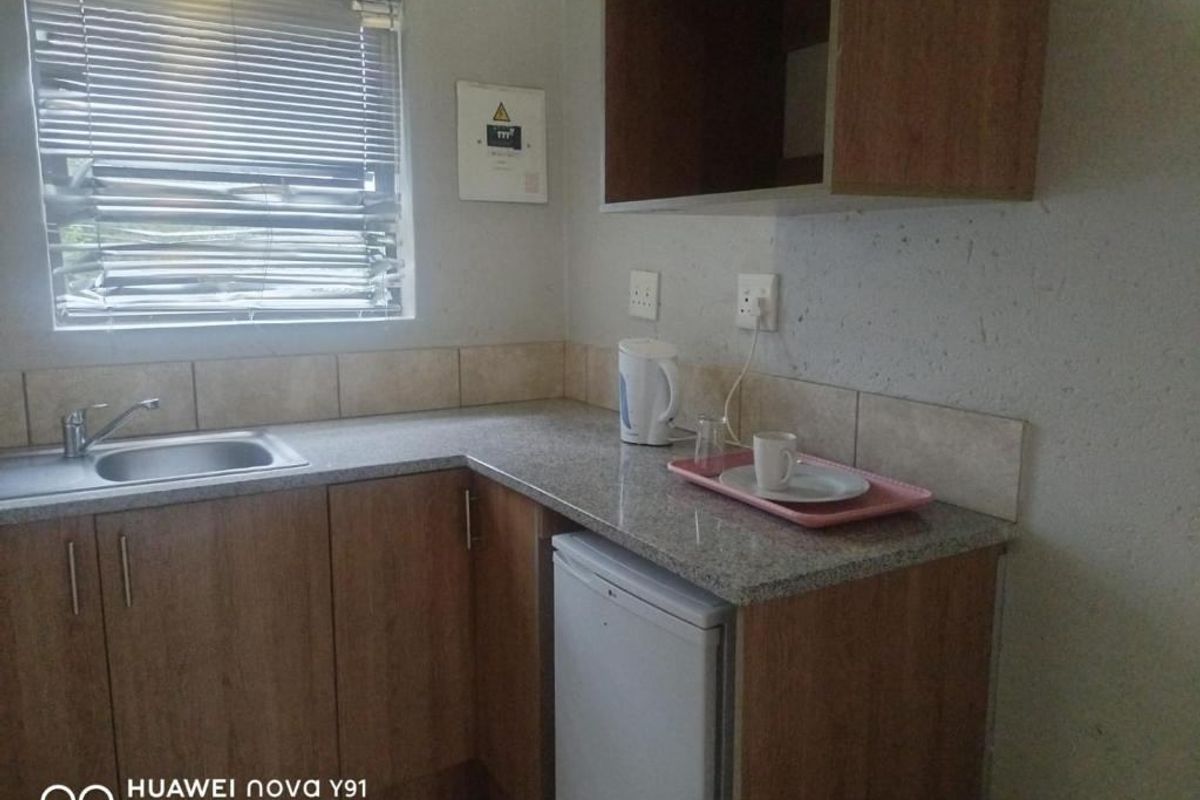 A compact kitchenette featuring wood-finish cabinetry, granite-look countertops, a stainless steel sink, and a window with fitted blinds.