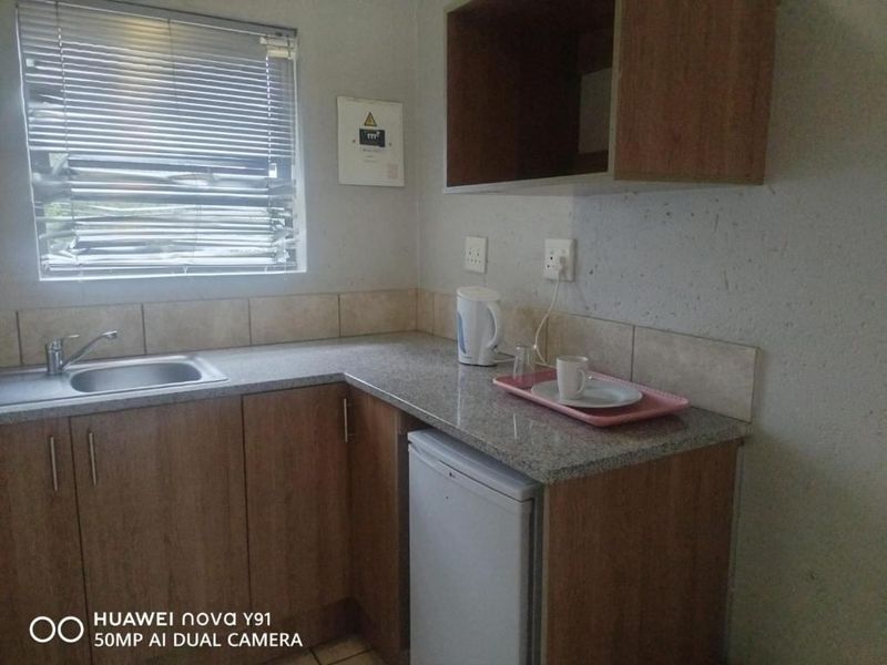 A compact kitchenette featuring wood-finish cabinetry, granite-look countertops, a stainless steel sink, and a window with fitted blinds.