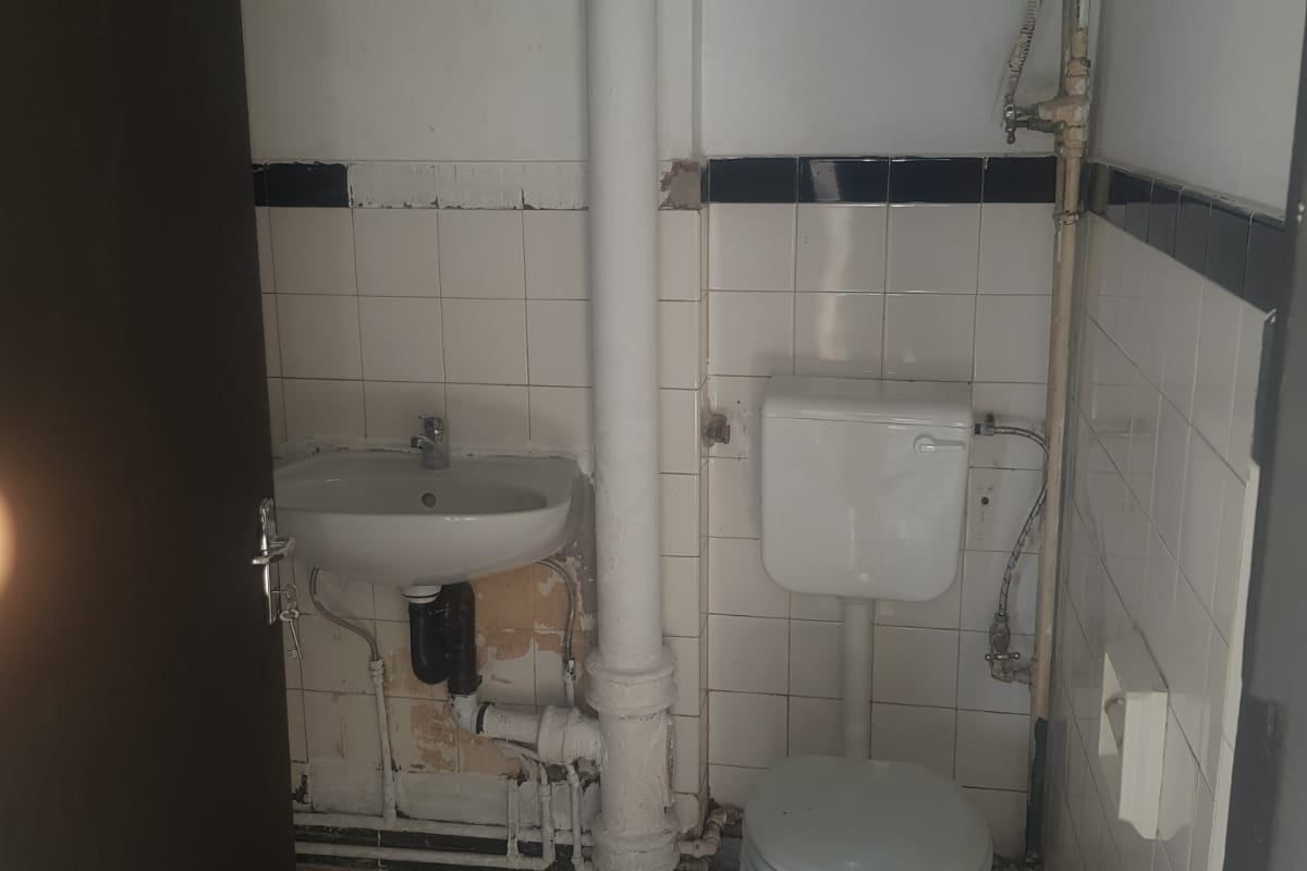 A compact bathroom featuring a standard toilet and wall-mounted basin, with exposed plumbing and dated tile finishes requiring renovation.