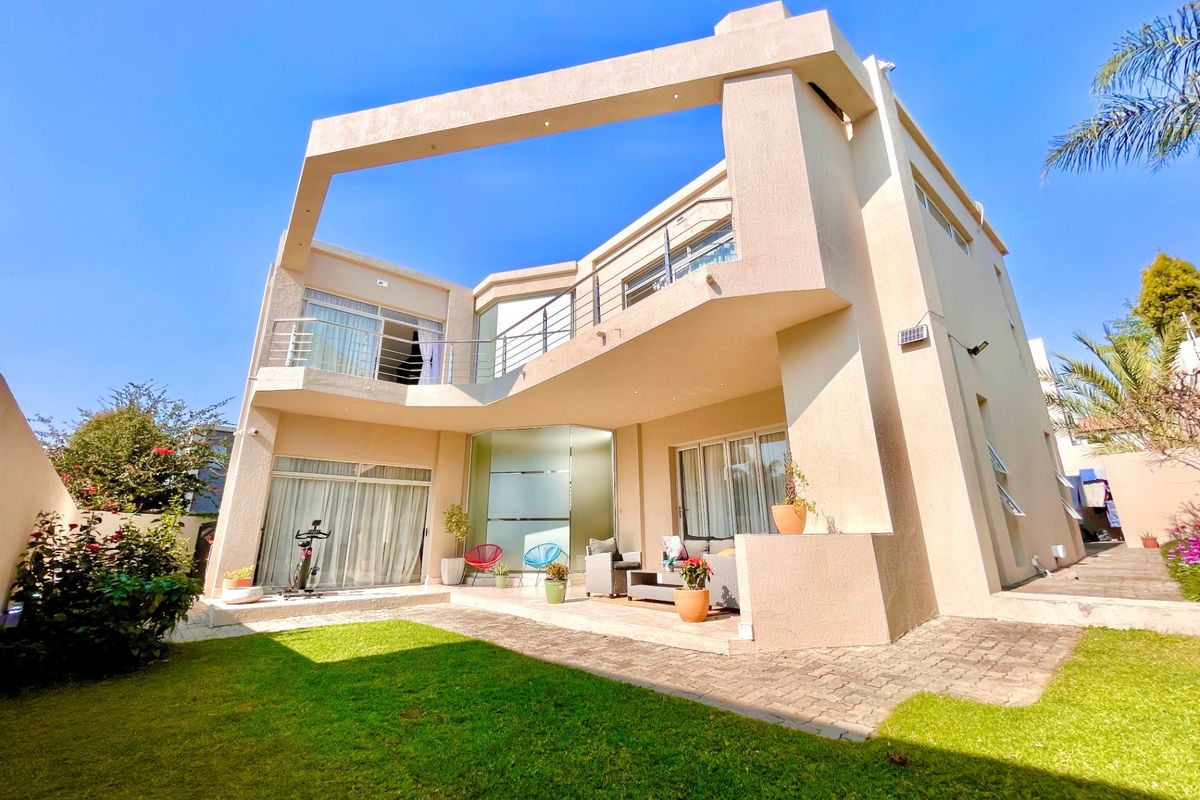 Rear view of a modern double-story residence featuring a paved patio, upper-level balcony with stainless steel railings, and a well-maintained garden.