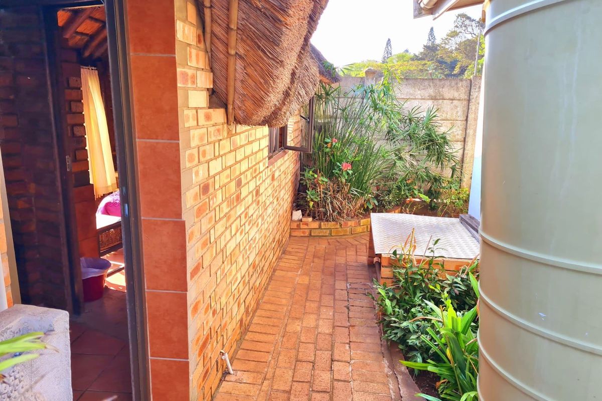 A side exterior view showing a brick-paved walkway, face brick wall with a thatch roof overhang, and a water storage tank.