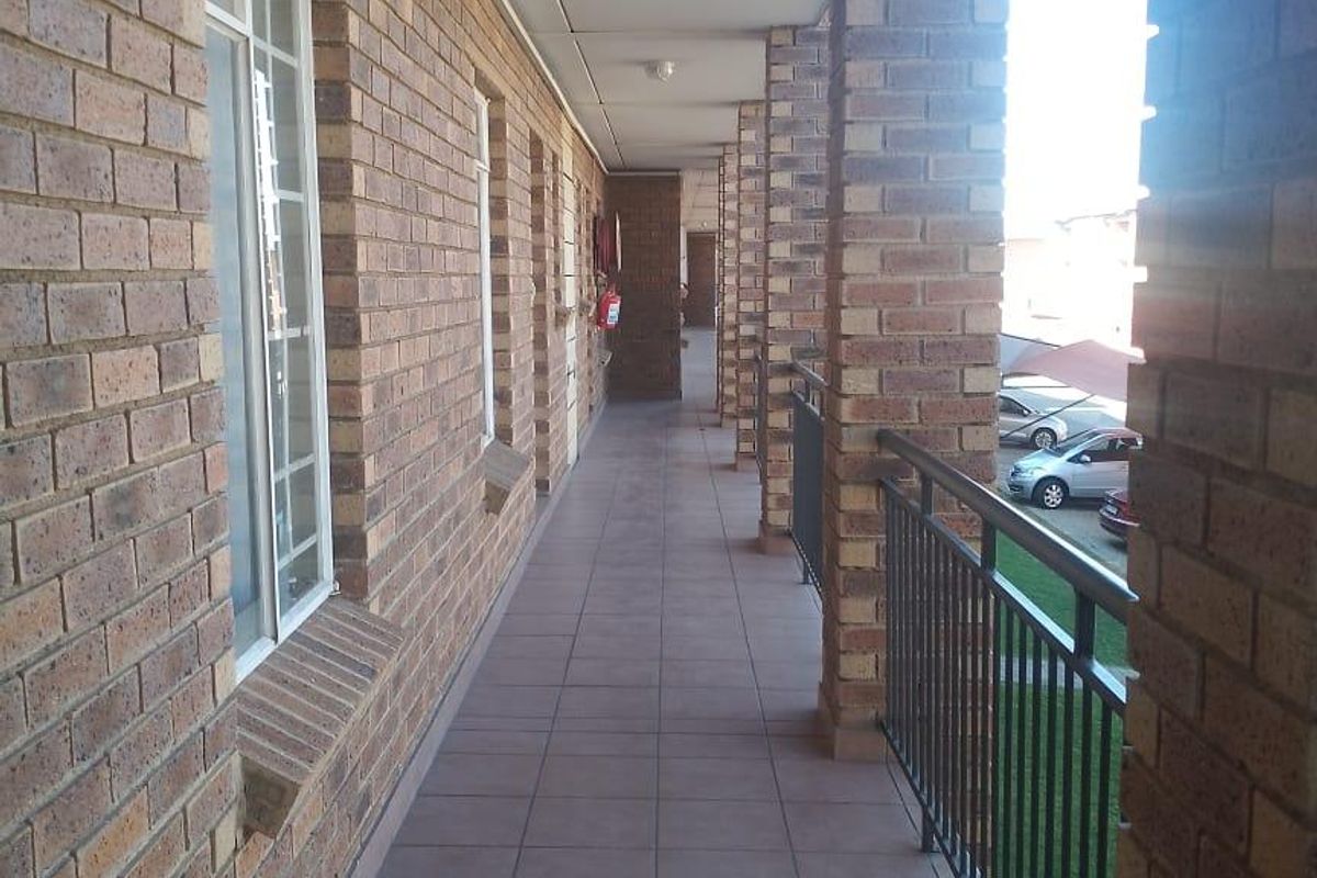 A covered outdoor balcony featuring brick walls, tiled flooring, and a metal balustrade overlooking the complex grounds.