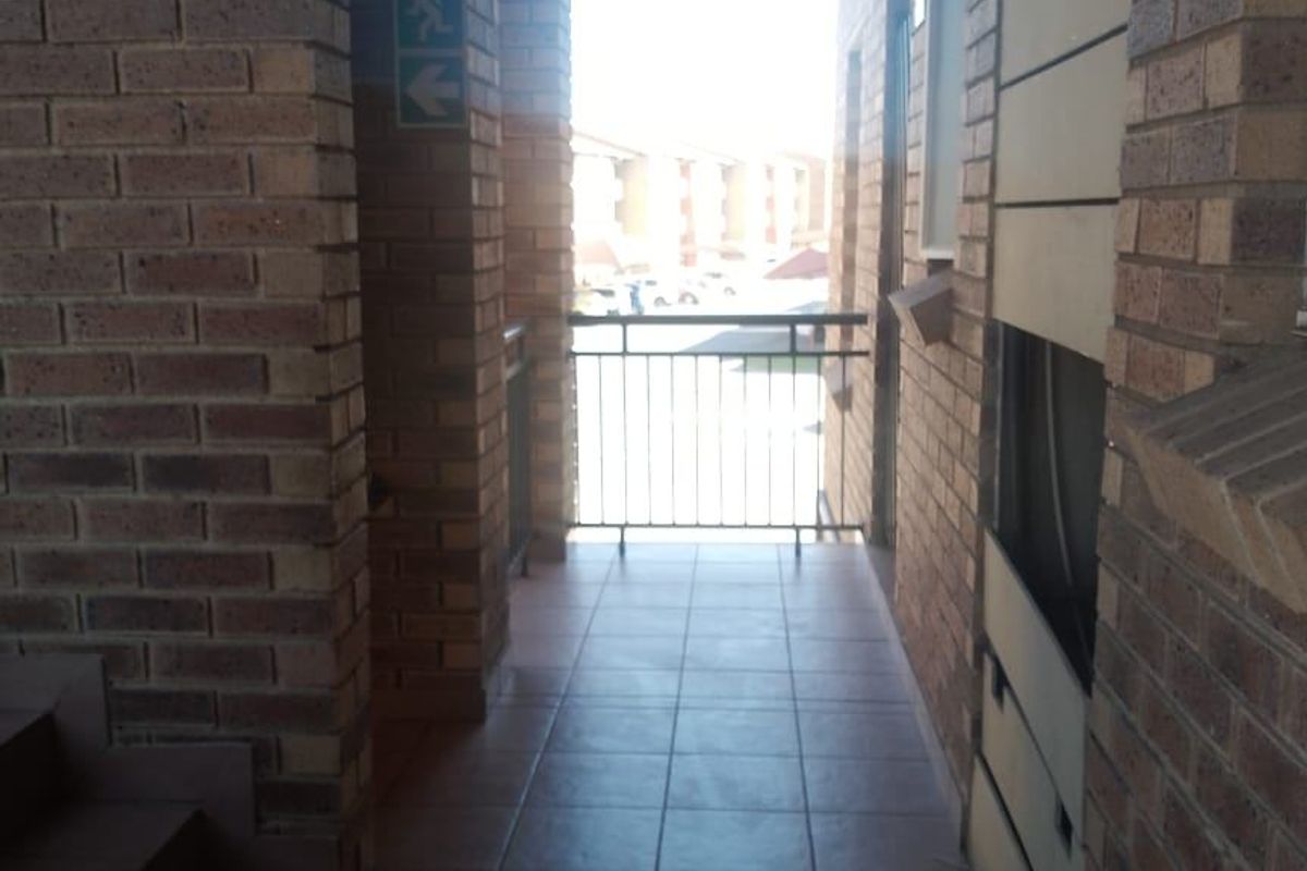 An outdoor-facing apartment hallway featuring exposed brick walls, tiled flooring, and a metal safety balustrade.