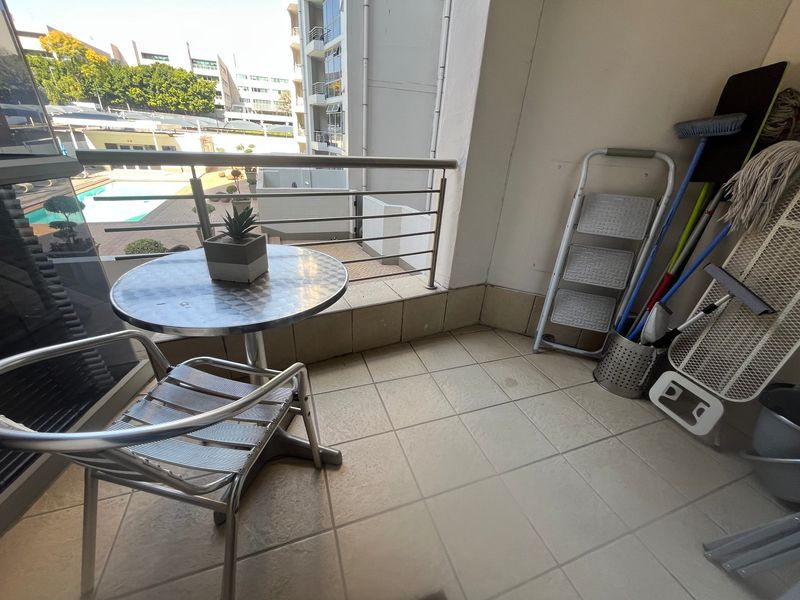 A compact balcony featuring tiled flooring and a stainless steel balustrade, offering an outdoor space with views of the complex pool area.