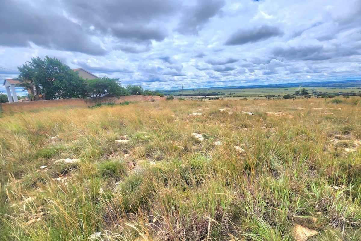 A wide-angle view of a vacant residential plot featuring natural grassland, rocky terrain, and distant landscape views under a cloudy sky.