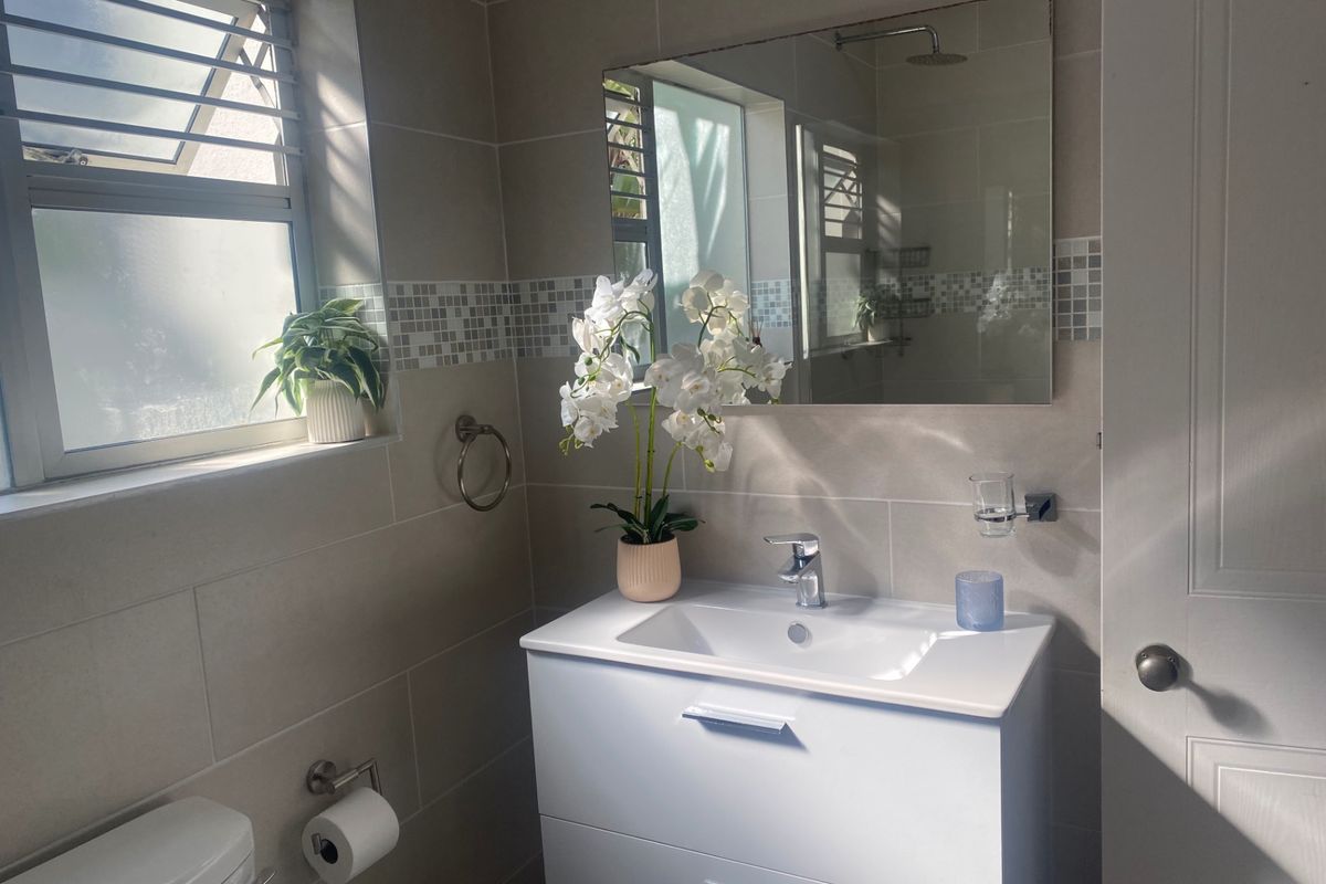 A modern bathroom featuring a white vanity cabinet with basin, wall-mounted mirror, tiled walls, and a louver window for natural ventilation.