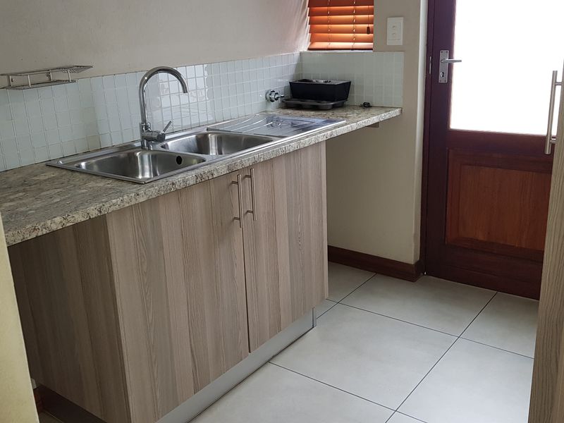A functional scullery area featuring laminate cabinetry, a stainless steel sink with mixer tap, tiled splashback, and direct access to the exterior.