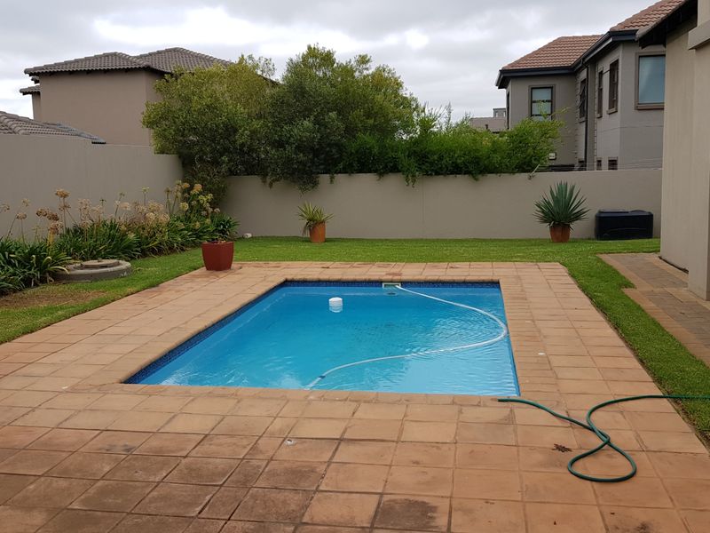 A private backyard featuring a swimming pool with a paved surround, lawn area, and perimeter boundary wall.