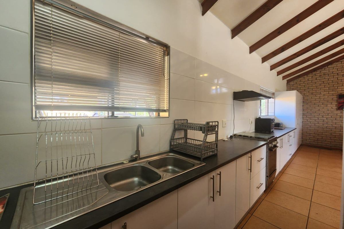 A functional kitchen featuring double stainless steel sinks, laminate countertops, exposed timber ceiling beams, and an exposed brick wall with tiled flooring.