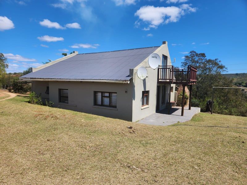 Rear view of the farm dwelling featuring a corrugated metal roof, a raised timber balcony, and a concrete patio area set within a grassy landscape.