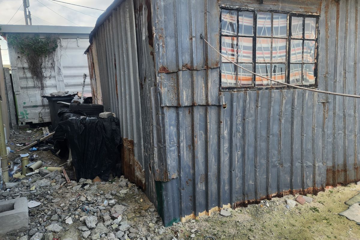 A view of a dilapidated structure featuring heavily rusted corrugated metal cladding and a weathered metal window frame on a vacant residential lot.