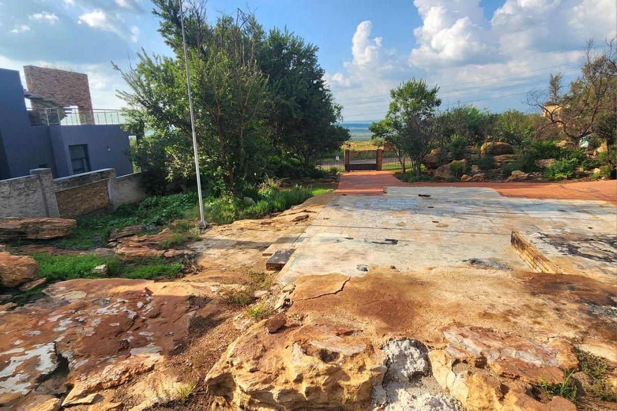 Rear exterior view of a residential plot featuring a concrete slab foundation, natural rock formations, and a paved pathway leading to a gated entrance.