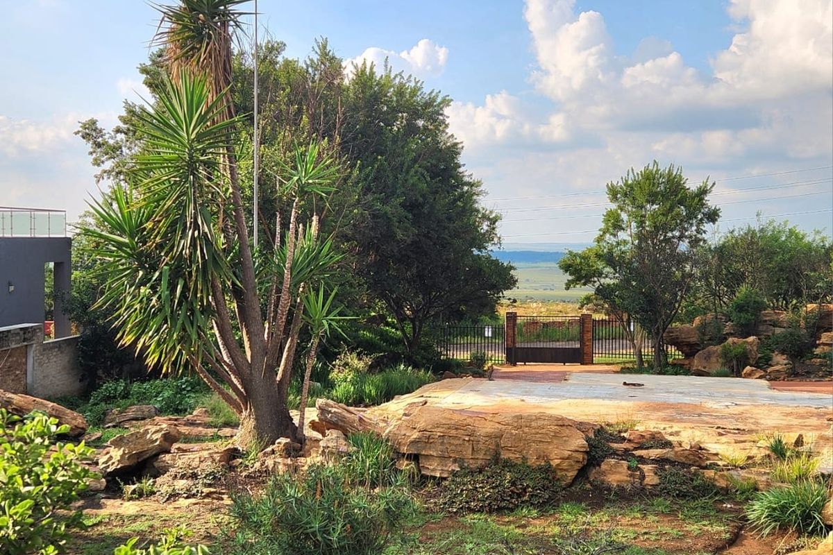 A landscaped residential garden featuring natural rock formations, mature trees, and a paved patio area with gated access to the surrounding landscape.