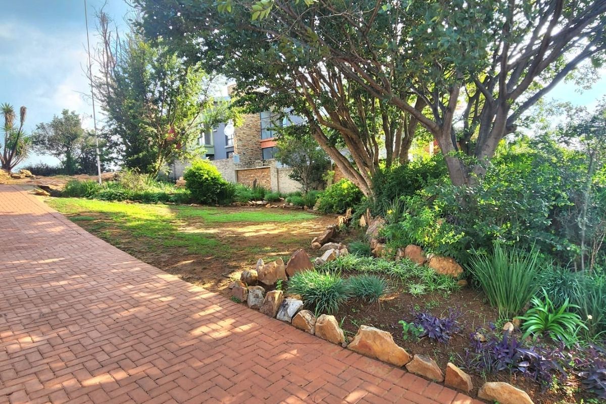 A well-maintained residential garden featuring a brick-paved driveway, mature trees, and stone-edged garden beds.