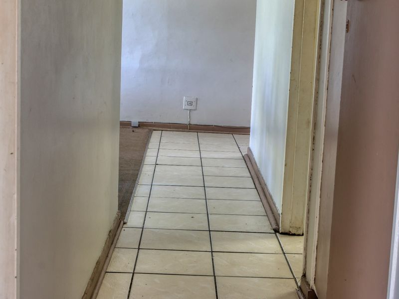 A narrow hallway featuring tiled flooring and visible wear on the skirting boards and wall finishes, leading into an adjacent carpeted room.