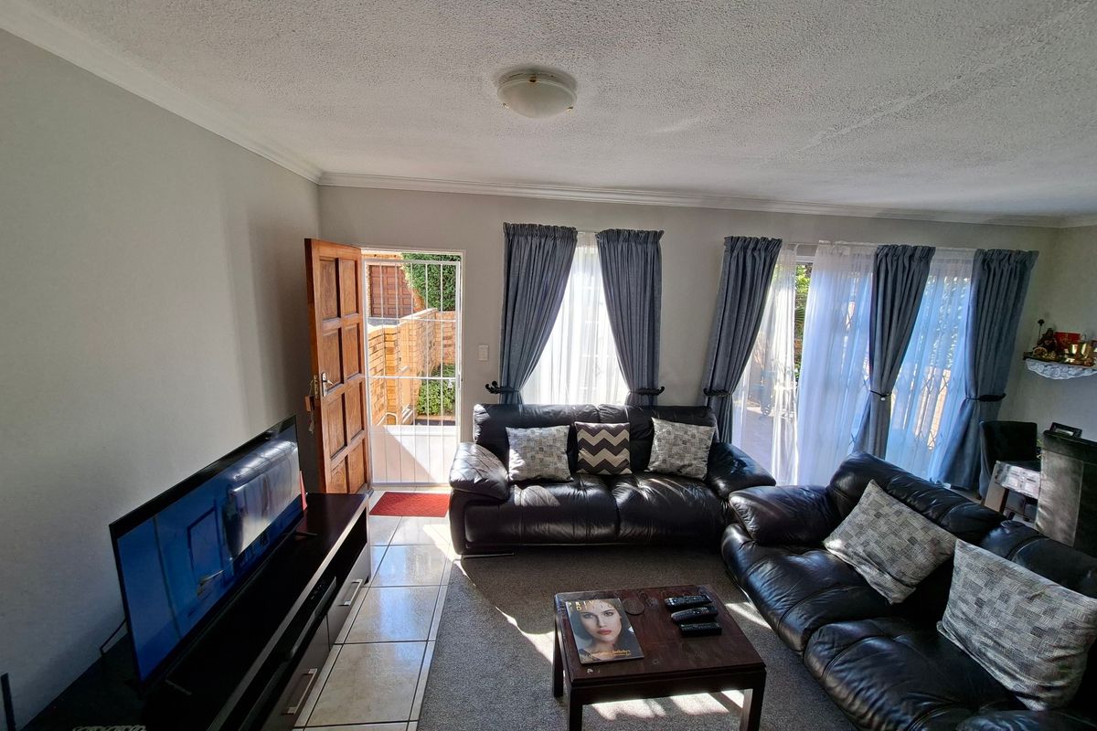 A living room featuring a mix of tiled and carpeted flooring, with direct access to an outdoor patio through a security gate and sliding doors.