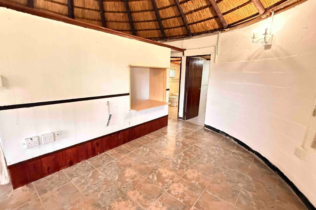 An empty bedroom featuring a traditional thatched roof, exposed timber rafters, and tiled flooring, with an adjoining bathroom visible through the doorway.