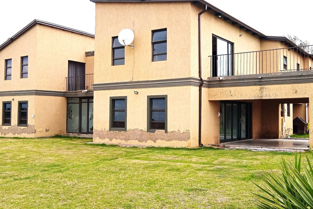 Rear view of a two-story residential property featuring a balcony, covered patio, and extensive lawn area, showing signs of exterior wall finish deterioration.