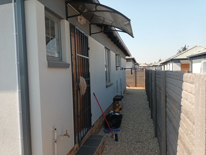 A side exterior view of the house featuring a security gate, protective awning, external tap, and a gravel pathway enclosed by a precast concrete wall.