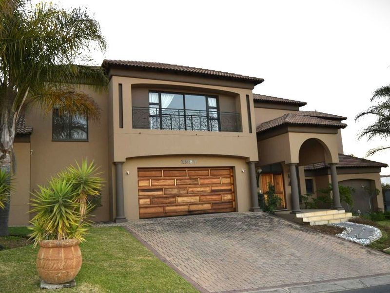 A well-maintained double-story house featuring a paved driveway, wooden garage door, arched entrance, and an upper-level balcony with wrought iron railings.