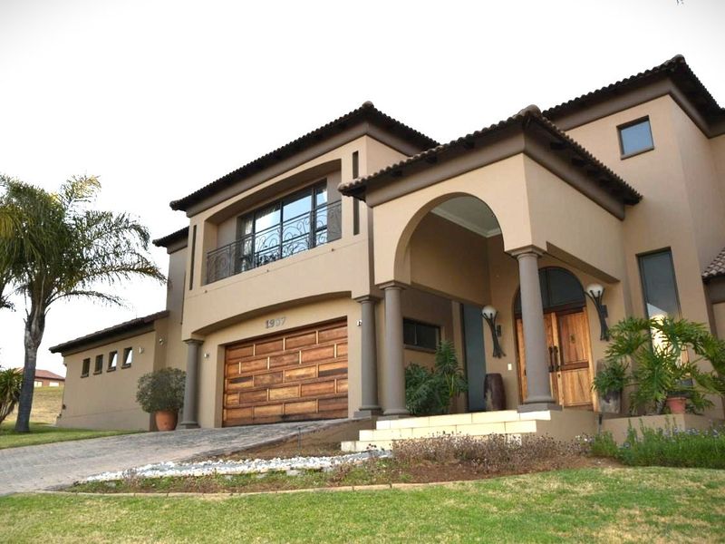 A well-maintained double-story residence featuring a paved driveway, wooden garage door, arched entrance porch with stone columns, and a balcony with wrought iron detailing.