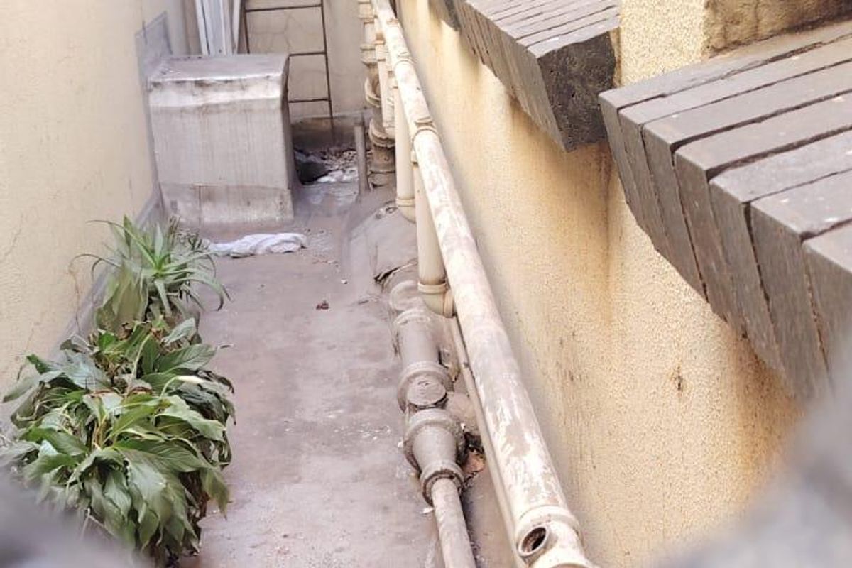 A narrow exterior rear service alley featuring exposed plumbing infrastructure and weathered wall finishes, viewed through a restricted opening.