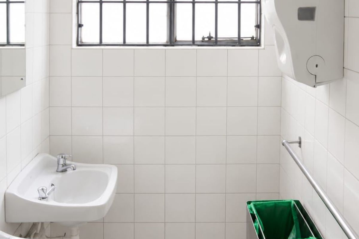 A functional commercial bathroom featuring dual pedestal washbasins, tiled surfaces, and a window with security bars.