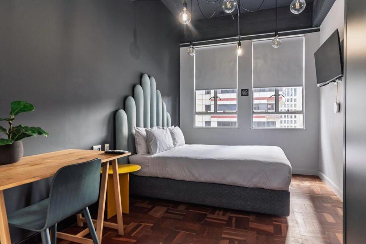 A modern bedroom featuring dark accent walls, parquet flooring, and large windows with roller blinds, complemented by industrial-style pendant lighting.