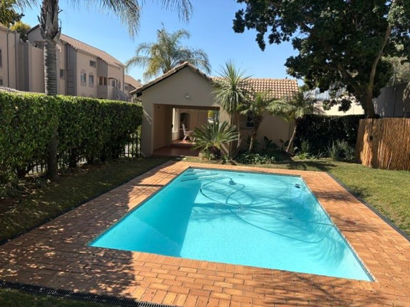 A well-maintained swimming pool area featuring a brick surround, landscaped garden, and an adjacent covered patio space.