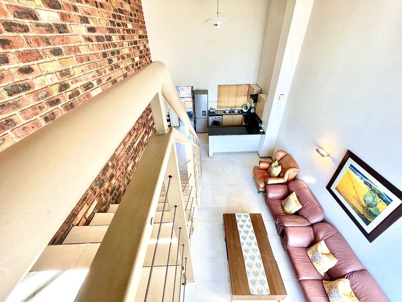 An elevated view of an open-plan living area featuring a double-volume space, exposed brick wall, staircase, and a functional kitchen with tiled flooring.