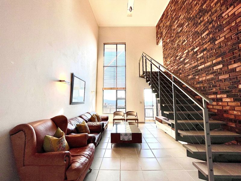 Spacious double-volume living area featuring an exposed brick feature wall, tiled flooring, and a modern staircase with metal balustrades.