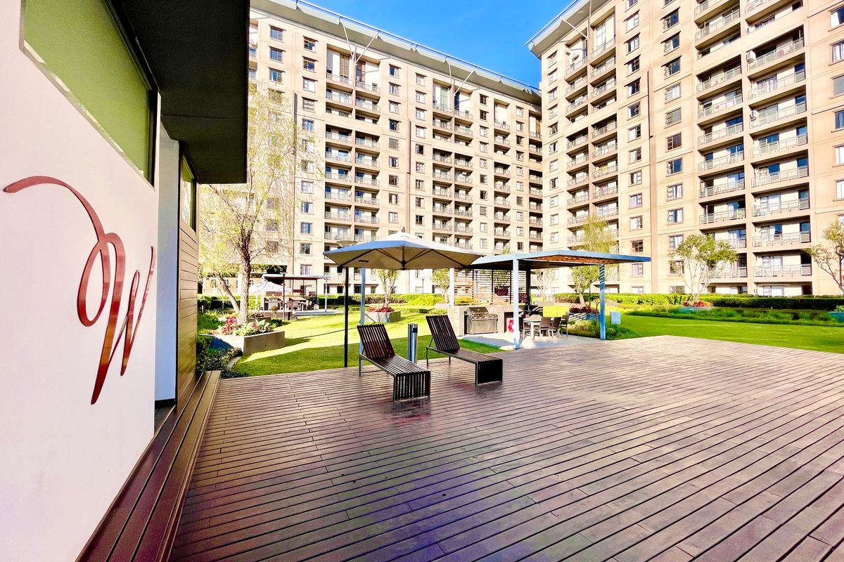 A well-maintained communal outdoor area featuring timber decking, landscaped gardens, and a built-in braai facility within a modern apartment complex.