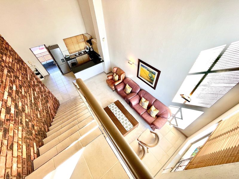 High-angle view of an open-plan living area featuring a double-volume ceiling, exposed brick feature wall, tiled floors, and an integrated kitchen.