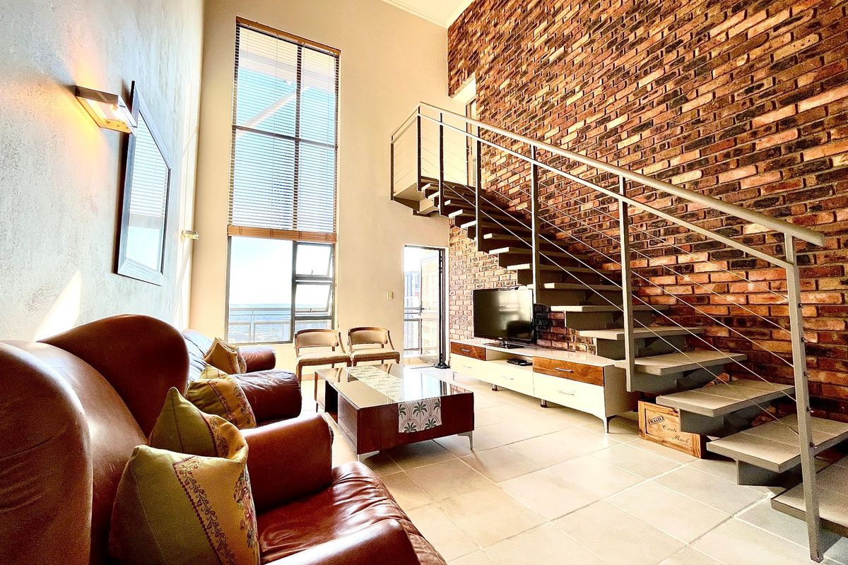 Spacious double-volume living area featuring an exposed brick feature wall, modern staircase, and large windows providing ample natural light.