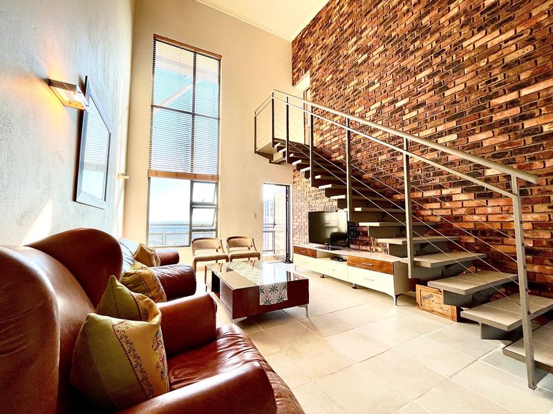 Spacious double-volume living area featuring an exposed brick feature wall, modern staircase, and large windows providing ample natural light.