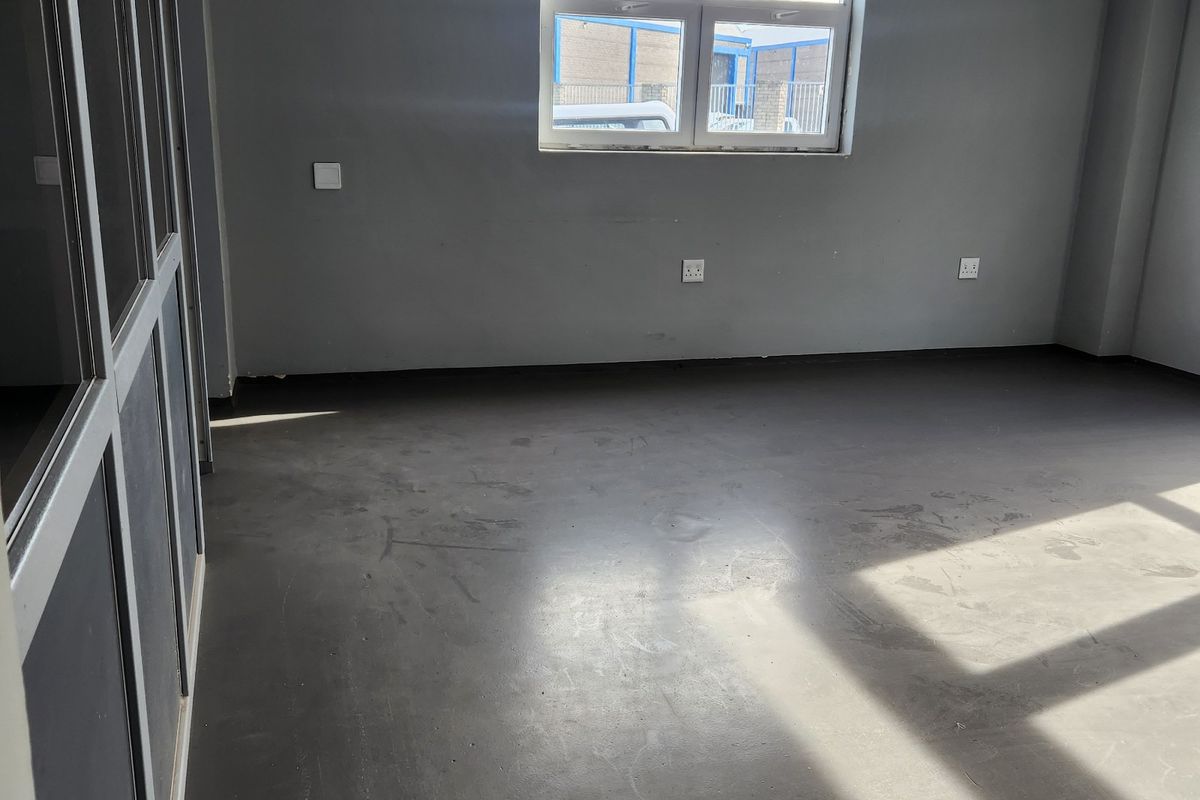 An empty commercial office space featuring grey flooring, an aluminium-framed glass partition, and natural light from a window.
