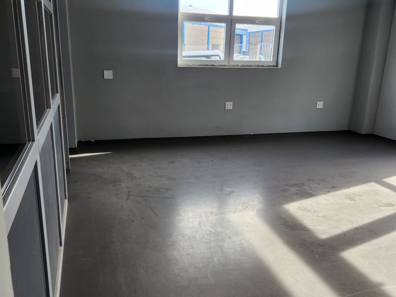 An empty commercial office space featuring grey flooring, an aluminium-framed glass partition, and natural light from a window.