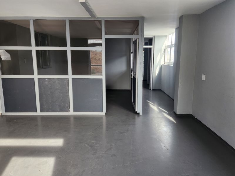 An empty commercial office space featuring grey vinyl flooring, a glass partition wall, and natural light from side windows.