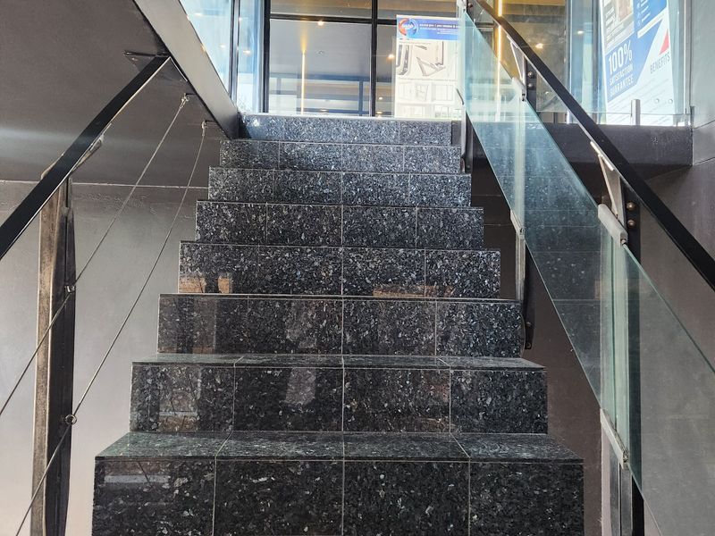 A modern commercial staircase featuring polished granite steps, a sleek glass balustrade, and a metal handrail, leading to an upper level.