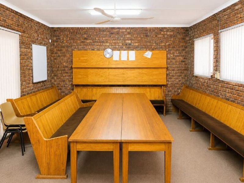 A functional multi-purpose room featuring exposed brick walls, a ceiling fan, and vertical blinds, suitable for a meeting or communal space.