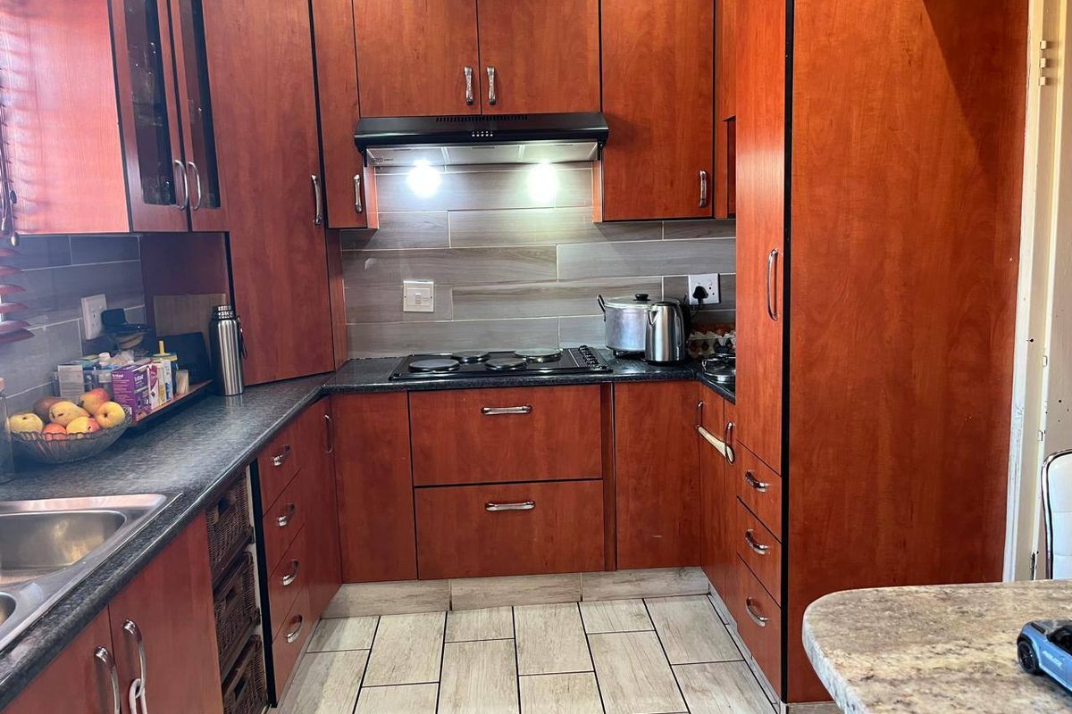 A functional kitchen featuring warm-toned wooden cabinetry, dark laminate countertops, a built-in electric hob with extractor fan, and modern tiled flooring.