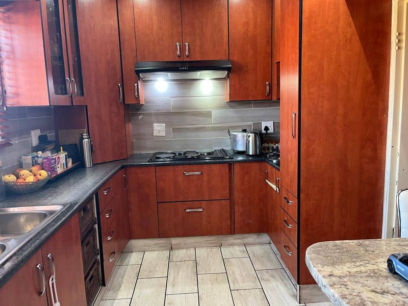 A functional kitchen featuring warm-toned wooden cabinetry, dark laminate countertops, a built-in electric hob with extractor fan, and modern tiled flooring.