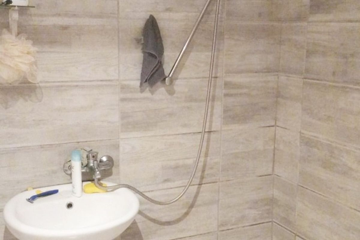 A compact bathroom featuring a pedestal basin and a wall-mounted handheld shower fixture with neutral ceramic tiling throughout.