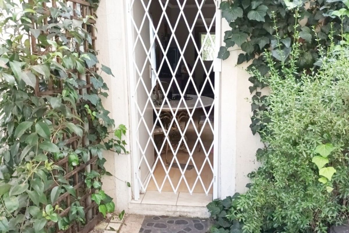 A rear entrance featuring a white security gate, surrounded by climbing ivy and established garden greenery with paved access.
