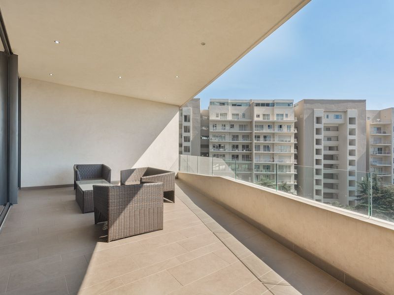 A spacious covered balcony featuring modern tiled flooring, a sleek glass balustrade, and recessed ceiling lighting, offering an open view of the surrounding apartment complex.